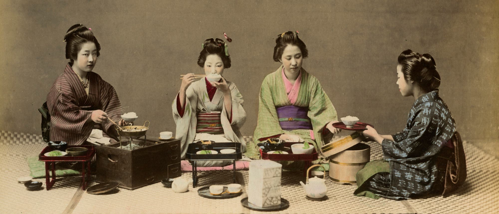 Vier Frauen in traditionellen japanischen Kimonos sitzen auf dem Boden und essen gemeinsam eine Mahlzeit. Sie sind um einen niedrigen Tisch versammelt, auf dem verschiedene Speisen und Teeservices arrangiert sind. Eine Frau h&auml;lt Essst&auml;bchen, w&auml;hrend eine andere eine Schale serviert. Die Szene vermittelt eine ruhige und gesellige Atmosph&auml;re. Im unteren rechten Eck ist der Text "25. Eating Rice" zu sehen.