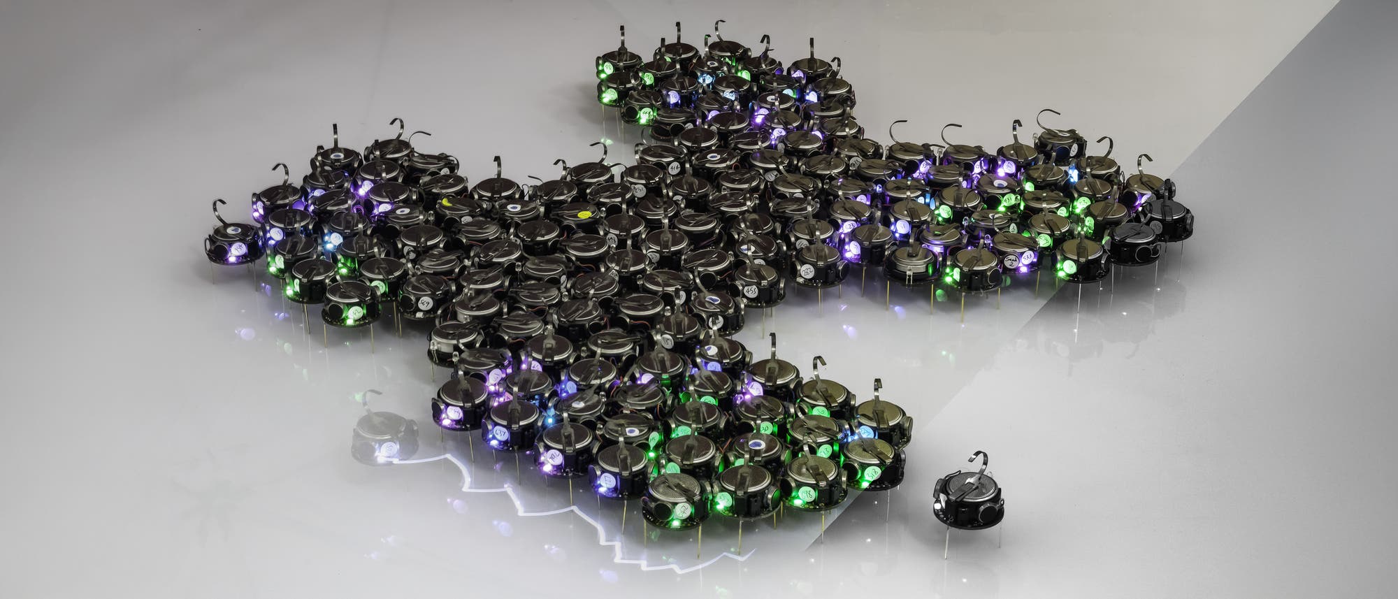 Eine Gruppe kleiner, runder Roboter mit leuchtenden LEDs in verschiedenen Farben (grün, blau, lila) formt eine dichte Formation auf einer glatten, reflektierenden Oberfläche. Ein einzelner Roboter ist etwas abseits von der Gruppe positioniert. Die Roboter sind gleichmäßig verteilt und scheinen miteinander zu interagieren, was auf eine koordinierte Bewegung oder ein gemeinsames Ziel hindeutet. Die Szene vermittelt ein Gefühl von Technologie und Zusammenarbeit.