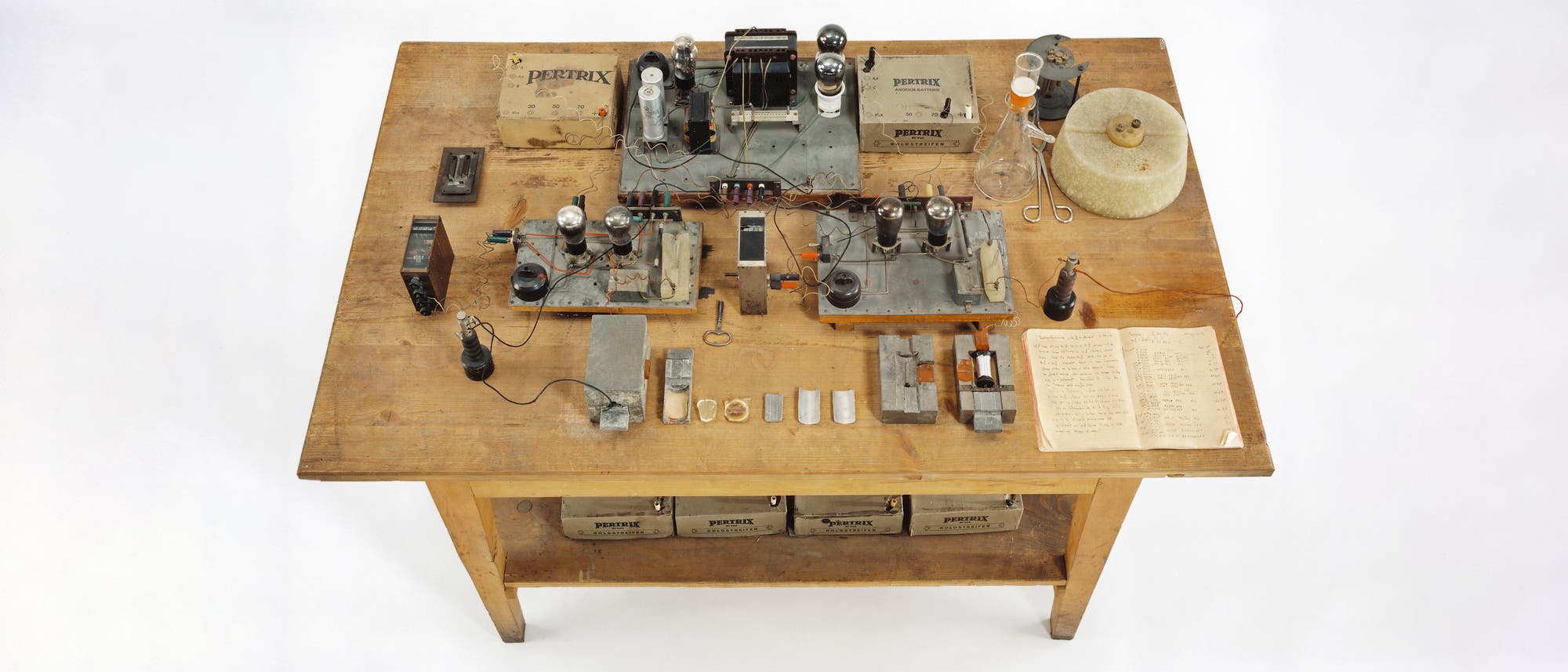 Ein Holztisch mit einer Anordnung von wissenschaftlichen Ger&auml;ten und Materialien. Auf dem Tisch befinden sich mehrere elektronische Komponenten, darunter R&ouml;hren, Kabel und Metallplatten. Rechts liegt ein offenes Notizbuch mit handschriftlichen Notizen. Im Hintergrund sind Glasflaschen und eine gro&szlig;e Spule zu sehen. Unter dem Tisch sind mehrere Batterien mit der Aufschrift "Pertrix" gelagert. Die Szene vermittelt den Eindruck eines experimentellen Labors.