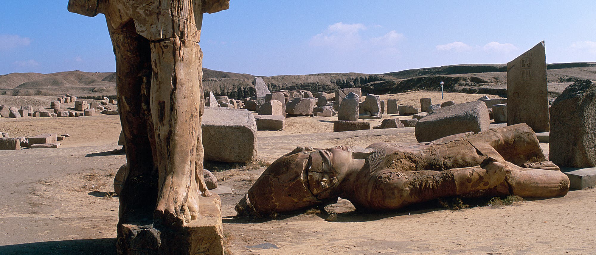 Eine antike arch&auml;ologische St&auml;tte mit &Uuml;berresten gro&szlig;er Steinskulpturen. Im Vordergrund steht ein besch&auml;digter Torso einer Statue, w&auml;hrend im Hintergrund eine liegende Statue und mehrere verstreute Steinelemente zu sehen sind. Der Himmel ist klar und blau, und die Umgebung ist eine trockene, sandige Landschaft.