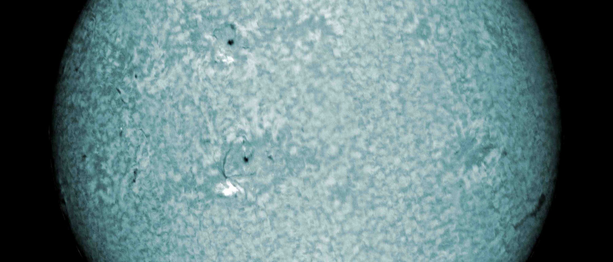 Eine wissenschaftliche Abbildung der Sonne, die in einem bl&auml;ulichen Farbton dargestellt ist. Die Oberfl&auml;che zeigt eine k&ouml;rnige Struktur mit dunkleren Flecken, die Sonnenflecken darstellen k&ouml;nnten. Diese Darstellung hebt die Textur und Aktivit&auml;t der Sonnenoberfl&auml;che hervor und bietet Einblicke in astronomische Ph&auml;nomene.