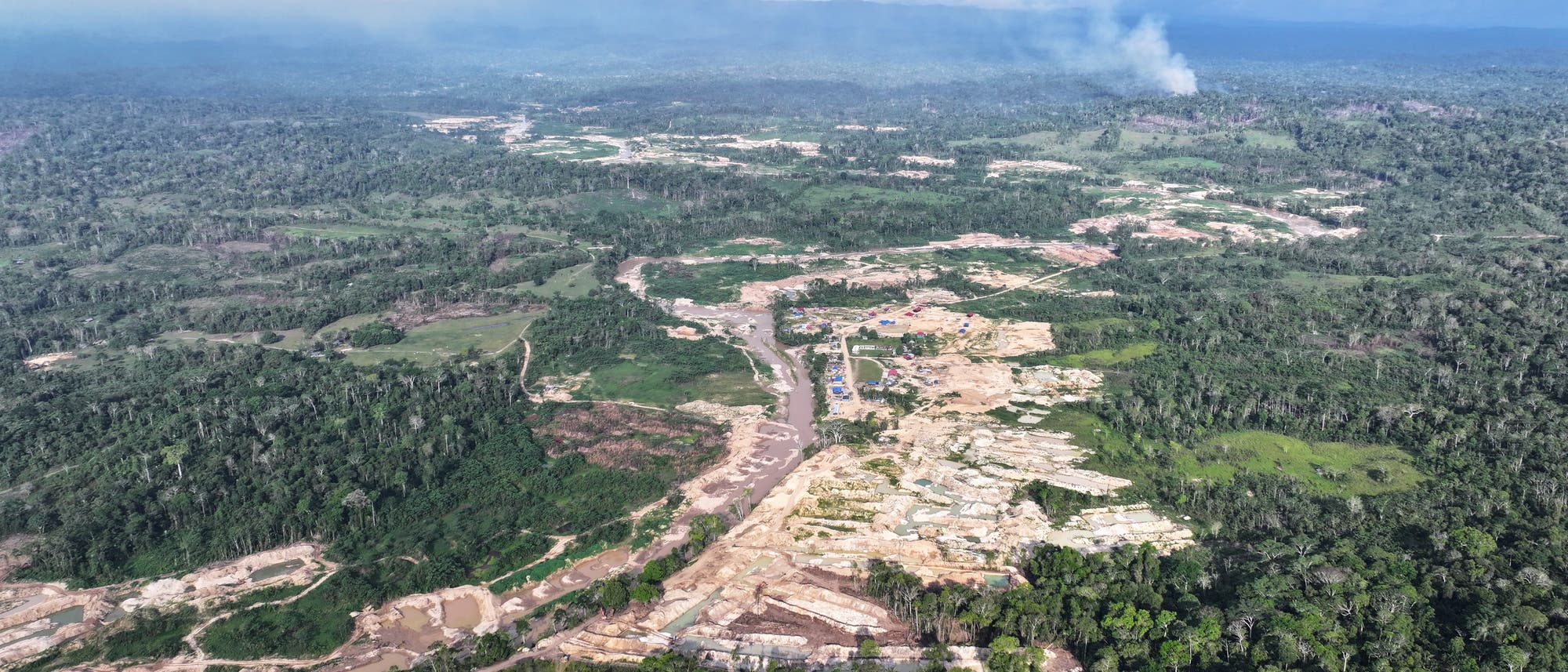 Luftaufnahme eines gro&szlig;en Waldgebiets mit deutlichen Spuren von Abholzung und Landnutzung. In der Mitte des Bildes sind mehrere helle Fl&auml;chen sichtbar, die auf menschliche Aktivit&auml;ten wie Bergbau oder Landwirtschaft hindeuten. Rauch steigt in der Ferne auf, was auf Brandrodung oder industrielle Prozesse hinweisen k&ouml;nnte. Der Himmel ist bew&ouml;lkt, und die Landschaft erstreckt sich bis zu den entfernten Bergen am Horizont.