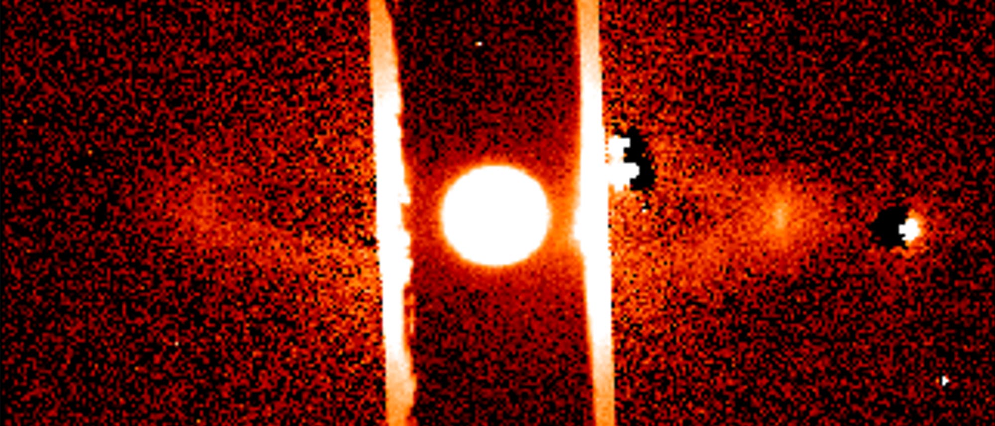 Ein astronomisches Bild zeigt ein helles, zentrales Objekt, vermutlich einen Stern, umgeben von einem r&ouml;tlichen Hintergrund. Zwei vertikale Streifen verlaufen parallel auf beiden Seiten des Objekts. Die Umgebung ist k&ouml;rnig, mit vereinzelten hellen Punkten, die m&ouml;glicherweise andere Himmelsk&ouml;rper darstellen. Das Bild vermittelt den Eindruck einer Weltraumaufnahme mit Fokus auf das zentrale Objekt.