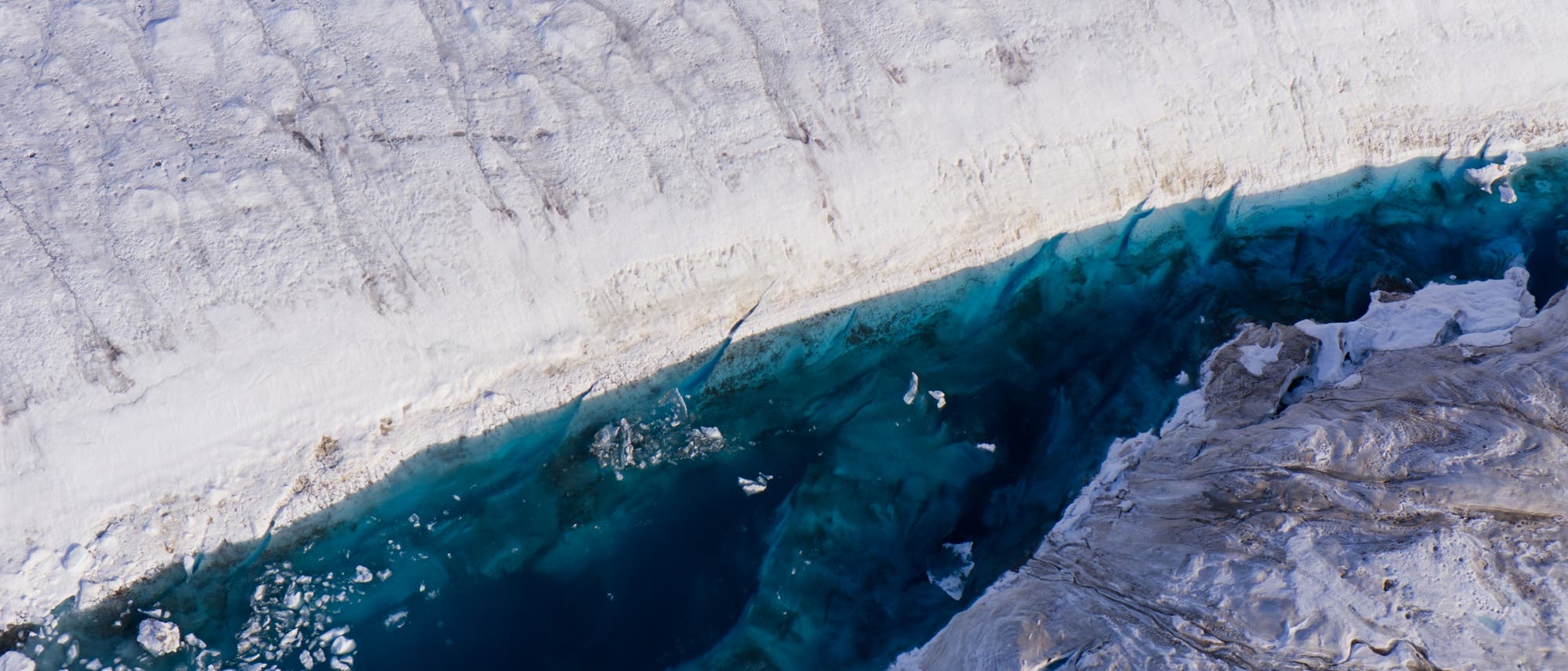 Luftaufnahme eines Gletschers mit schneebedeckter Oberfläche und einer tiefblauen Schmelzwasserlagune. Die weiße Schneedecke kontrastiert mit dem klaren, blauen Wasser, das von Eisschollen durchzogen ist. Die Struktur des Eises und die Farbverläufe im Wasser sind deutlich erkennbar.