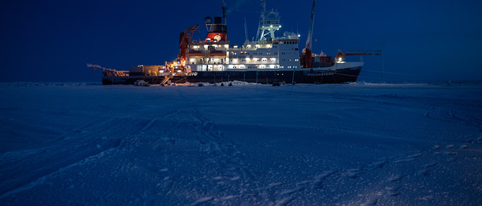 Ein gro&szlig;es Forschungsschiff, beleuchtet in der Dunkelheit, liegt im gefrorenen arktischen Eis. Der Nachthimmel ist tiefblau, und das Schiff strahlt warmes Licht aus, das die Umgebung erhellt. Im Vordergrund sind Spuren im Schnee zu sehen, die zum Schiff f&uuml;hren. Das Bild vermittelt eine ruhige, aber eindrucksvolle Atmosph&auml;re der Polarregion.