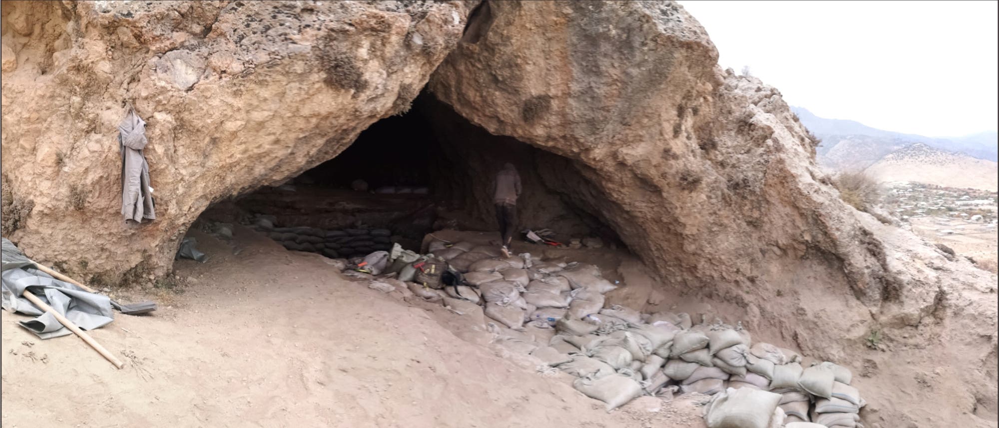 Eine große Höhle in einer felsigen Landschaft, deren Eingang mit Sandsäcken gesichert ist. Im Inneren der Höhle sind weitere Sandsäcke sichtbar. Eine Person steht in der Höhle, umgeben von Ausrüstungsgegenständen. Die Umgebung ist trocken und bergig, mit einem weiten Blick auf die umliegende Landschaft.