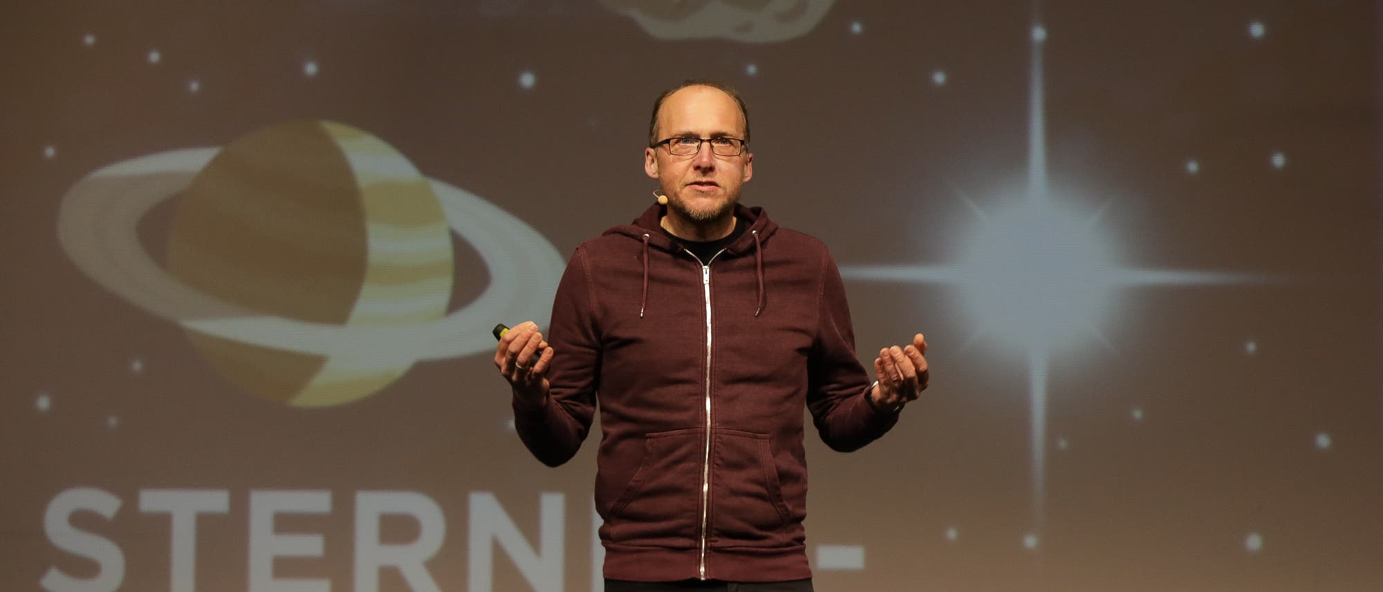 Florian Freistetter hält einen Vortrag vor einer projizierten Illustration eines Planeten und einem leuchtenden Stern. Im Hintergrund ist in großen Buchstaben der Name seines Podcasts, "STERNENGESCHICHTEN", zu lesen. Er trägt einen dunklen Hoodie und gestikuliert. Florian Freistetter hält einen Vortrag vor einer projizierten Illustration eines Planeten und einem leuchtenden Stern. Im Hintergrund ist in großen Buchstaben der Name seines Podcasts, "STERNENGESCHICHTEN", zu lesen. Er trägt einen dunklen Hoodie und gestikuliert.