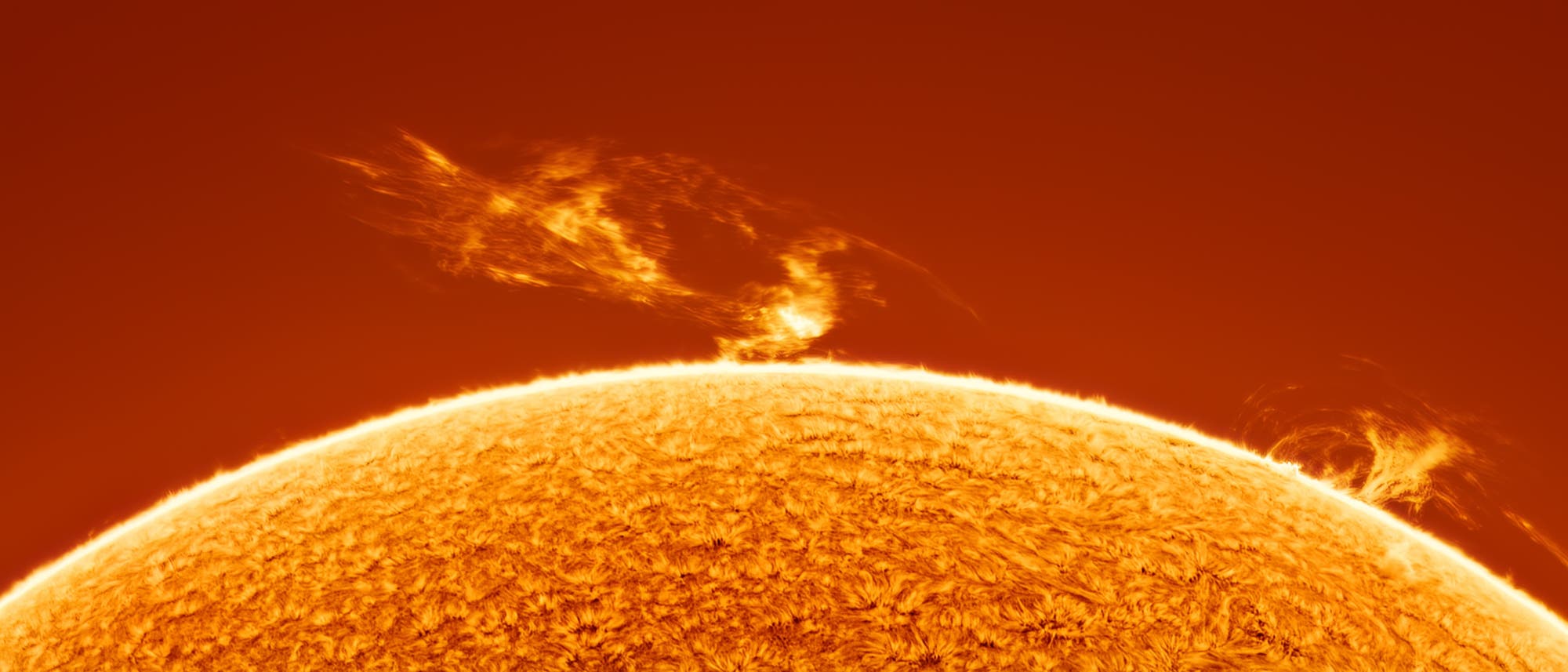 Eine Nahaufnahme der Sonnenoberfläche zeigt eine leuchtende, orangefarbene Textur mit zwei auffälligen Sonnenprotuberanzen, die sich in den Weltraum erstrecken. Die Protuberanzen erscheinen als helle, flammenartige Strukturen, die sich von der Sonnenoberfläche abheben. Der Hintergrund ist ein tiefes Orange, das die Intensität der Sonnenaktivität unterstreicht.