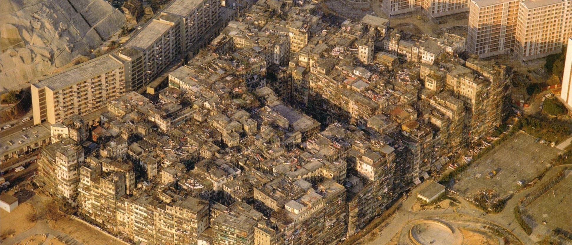 Luftaufnahme einer dichten städtischen Siedlung, umgeben von moderneren Hochhäusern und offenen Flächen. Die zentrale Struktur besteht aus eng aneinandergebauten, verwitterten Gebäuden, die ein labyrinthisches Muster bilden. Im Vordergrund sind geschwungene Wege und ein Parkplatz zu sehen. Die Szene zeigt den Kontrast zwischen alter und neuer Architektur.