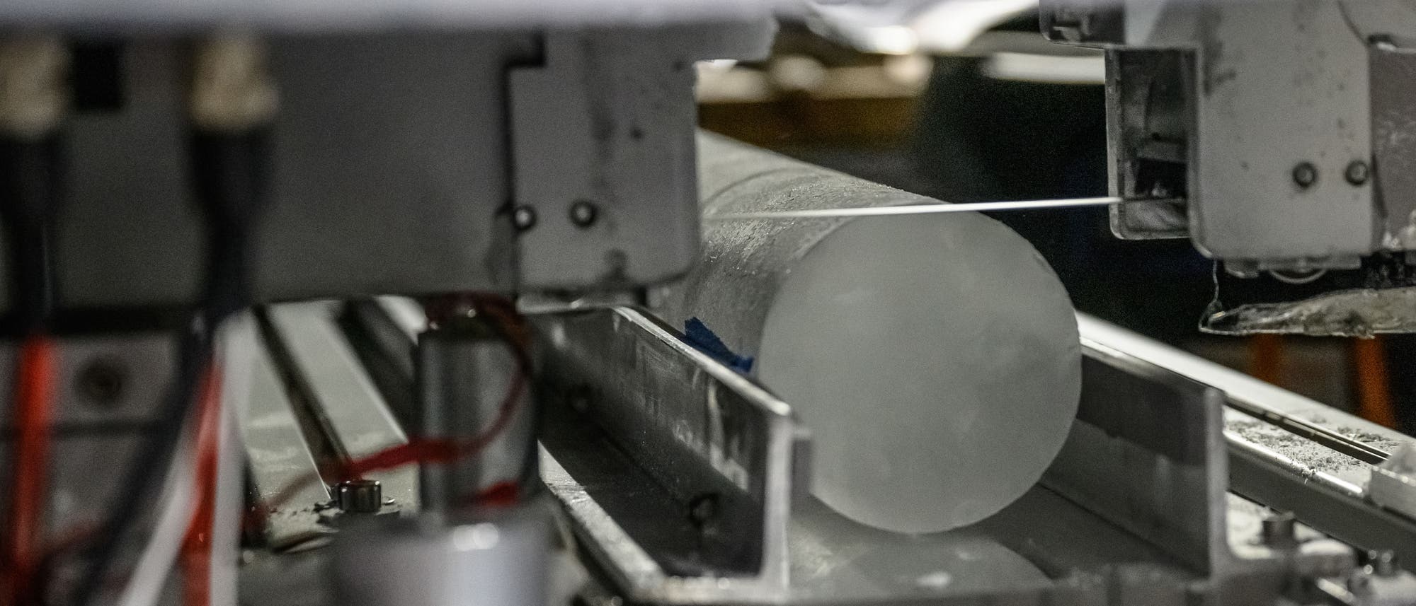 Eine Maschine schneidet einen zylindrischen Eisbohrkern. Der Eisbohrkern liegt in einer metallischen Halterung, während ein dünner Draht ihn durchtrennt. Die Umgebung wirkt industriell und technisch. Eine Maschine schneidet einen zylindrischen Eisbohrkern. Der Eisbohrkern liegt in einer metallischen Halterung, während ein dünner Draht ihn durchtrennt. Die Umgebung wirkt industriell und technisch.
