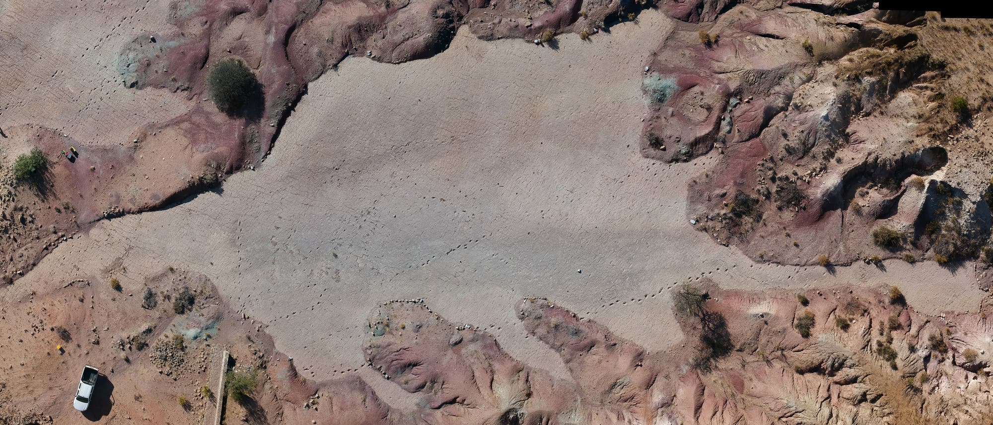 Luftaufnahme einer trockenen, felsigen Landschaft mit rötlichen und bräunlichen Tönen. In der unteren linken Ecke ist ein weißes Fahrzeug zu sehen. Die Oberfläche zeigt Spuren von Theropoden.