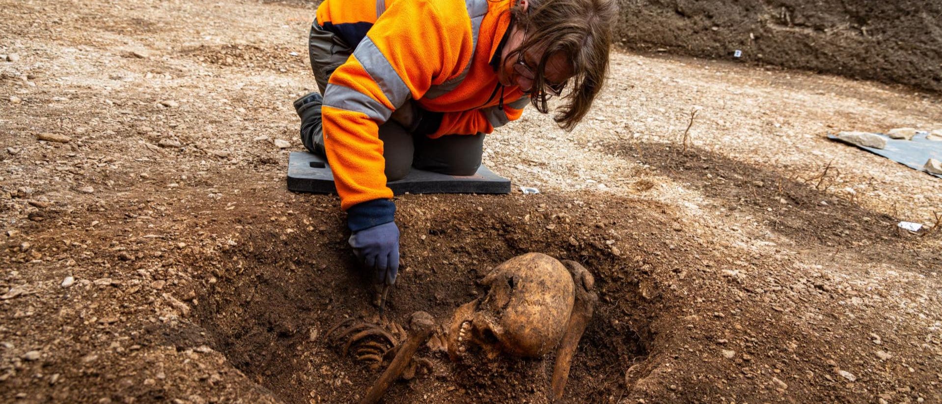 Eine Arch&auml;ologin in orangefarbener Arbeitskleidung kniet auf einer Ausgrabungsst&auml;tte und untersucht ein freigelegtes menschliches Skelett im Boden. Im Hintergrund sind weitere Arbeiter und Geb&auml;ude zu sehen. Die Szene zeigt eine arch&auml;ologische Untersuchung in einem urbanen Umfeld.