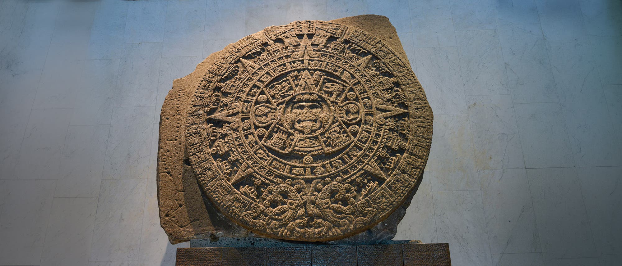 Steinrelief mit komplexen Gravuren, das ein kreisförmiges Design zeigt, das an einen antiken Kalender erinnert. Das Relief ist reich an Symbolen und Mustern, die radial angeordnet sind. Es befindet sich in einem Museum auf einem Sockel und ist von einer neutralen Wand umgeben. Die kunstvolle Darstellung spiegelt die kulturelle Bedeutung und das handwerkliche Können der Zivilisation wider, die es geschaffen hat. Steinrelief mit komplexen Gravuren, das ein kreisförmiges Design zeigt, das an einen antiken Kalender erinnert. Das Relief ist reich an Symbolen und Mustern, die radial angeordnet sind. Es befindet sich in einem Museum auf einem Sockel und ist von einer neutralen Wand umgeben. Die kunstvolle Darstellung spiegelt die kulturelle Bedeutung und das handwerkliche Können der Zivilisation wider, die es geschaffen hat.