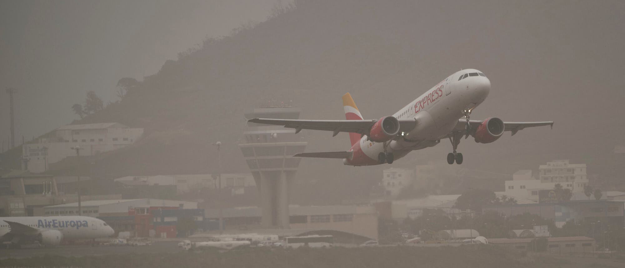 Ein Flugzeug hebt von einer Landebahn ab, w&auml;hrend im Hintergrund ein Kontrollturm und mehrere Geb&auml;ude zu sehen sind. Der Himmel ist bew&ouml;lkt und die Sicht ist leicht eingeschr&auml;nkt. Auf dem Flugzeug ist der Schriftzug "Iberia Express" zu erkennen. Ein weiteres Flugzeug der "Air Europa" steht am Boden. Die Szene spielt sich auf einem Flughafen ab, umgeben von H&uuml;geln.