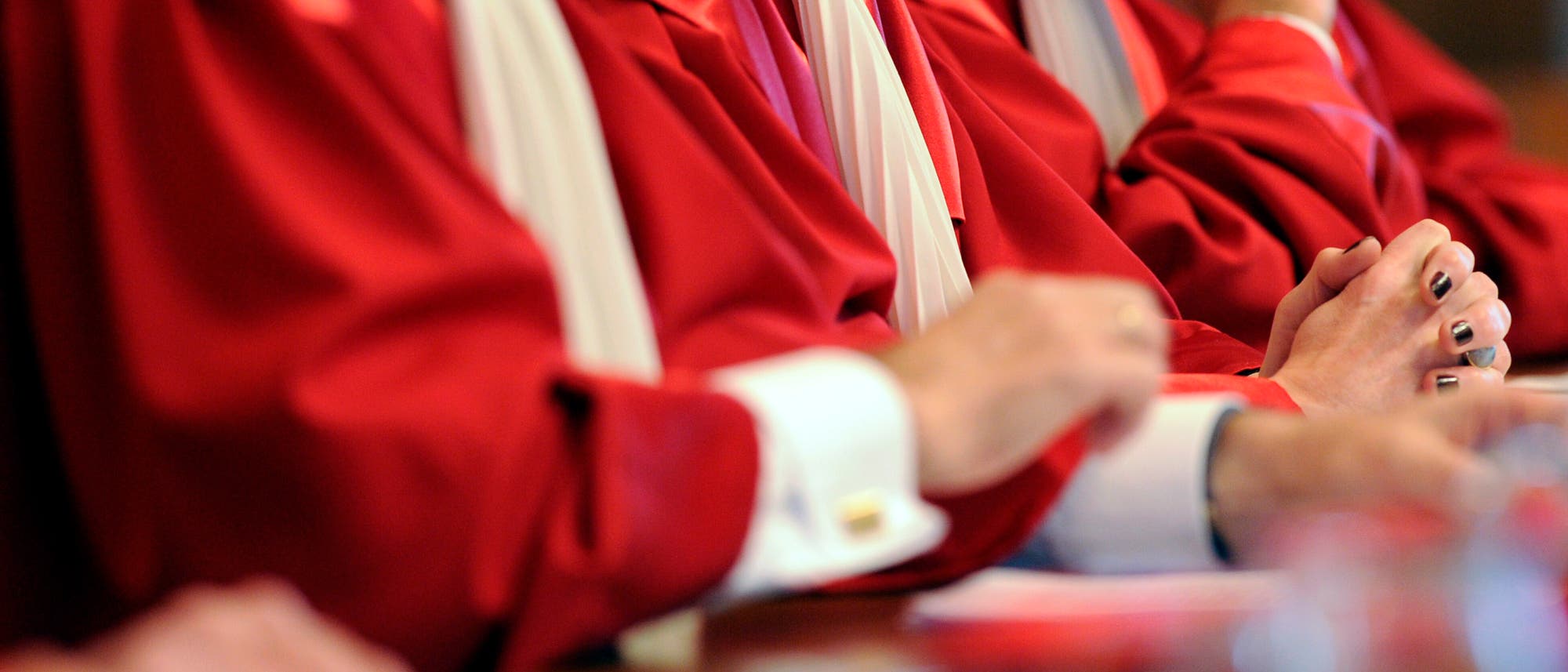 Eine Gruppe von Richterinnen und Richtern in roten Roben sitzt nebeneinander an einem Tisch. Die Gesichter sind nicht zu sehen, da das Bild nach oben abgeschnitten ist. Die Hände der Personen sind gefaltet oder ruhen auf dem Tisch. Die Roben haben weiße Kragen. Die Szene vermittelt einen formellen, gerichtlichen oder zeremoniellen Kontext.