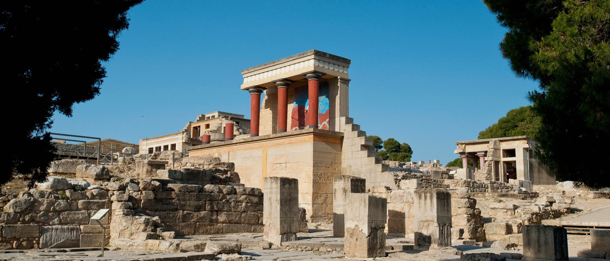 Ruinen des Palastes von Knossos auf Kreta, Griechenland, bei klarem Himmel. Im Vordergrund sind antike Steinstrukturen und S&auml;ulen zu sehen, w&auml;hrend im Hintergrund ein restaurierter Teil des Palastes mit roten S&auml;ulen und Wandmalereien hervorsticht. B&auml;ume rahmen die Szene ein.