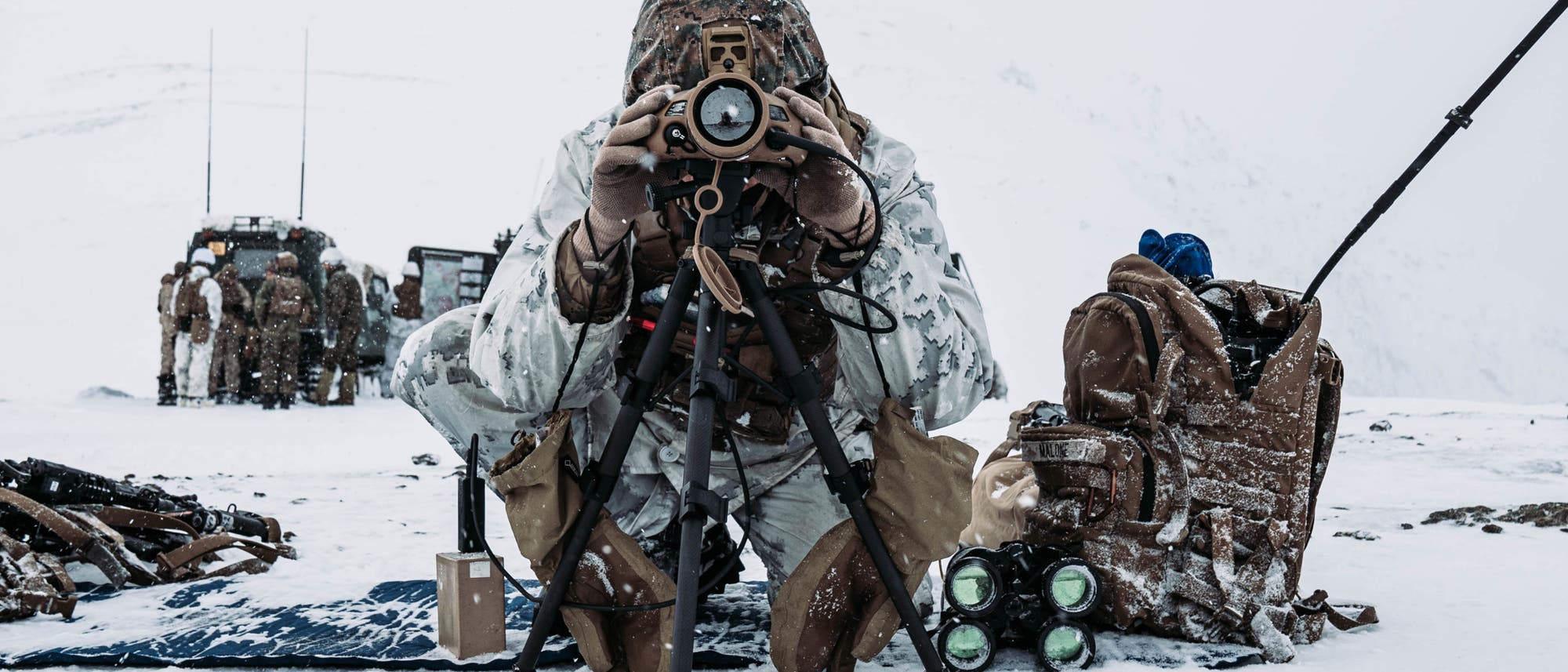 Eine Person in Tarnkleidung kniet im Schnee und blickt durch ein Fernglas, das auf einem Stativ montiert ist. Im Hintergrund sind weitere Personen und Ausr&uuml;stungsgegenst&auml;nde zu sehen. Die Szene spielt in einer verschneiten, abgelegenen Umgebung. Die Ausr&uuml;stung umfasst Taschen und technische Ger&auml;te, die auf dem Boden verteilt sind.