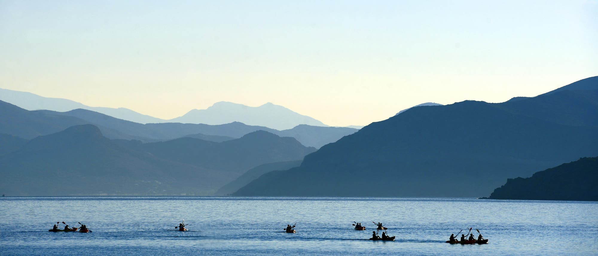 Mehrere Kanus mit Menschen paddeln auf einem ruhigen See, umgeben von einer beeindruckenden Berglandschaft im Hintergrund. Die Szene vermittelt Ruhe und Naturverbundenheit. Die Silhouetten der Berge sind im sanften Licht der Dämmerung zu sehen. Mehrere Kanus mit Menschen paddeln auf einem ruhigen See, umgeben von einer beeindruckenden Berglandschaft im Hintergrund. Die Szene vermittelt Ruhe und Naturverbundenheit. Die Silhouetten der Berge sind im sanften Licht der Dämmerung zu sehen.