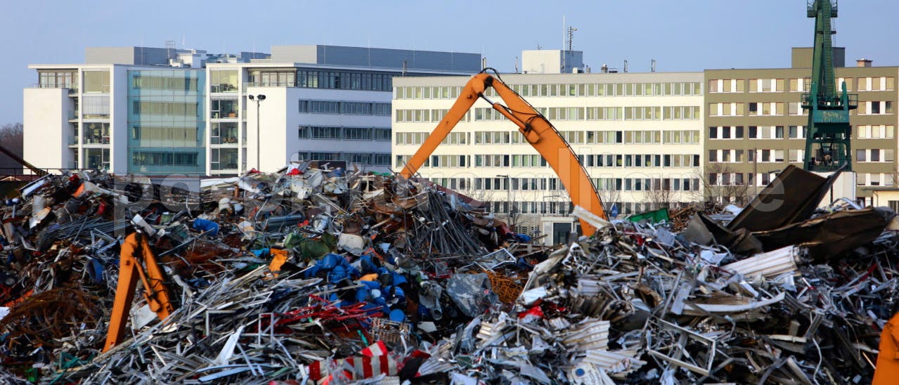 Ein großer Schrottplatz mit einem Haufen aus Metallabfällen, darunter bunte Teile und Schrottstücke. Zwei orangefarbene Kräne sind im Einsatz, um den Schrott zu bewegen. Im Hintergrund sind moderne Bürogebäude zu sehen, die den städtischen Charakter der Umgebung unterstreichen. Der Himmel ist klar und blau. Ein großer Schrottplatz mit einem Haufen aus Metallabfällen, darunter bunte Teile und Schrottstücke. Zwei orangefarbene Kräne sind im Einsatz, um den Schrott zu bewegen. Im Hintergrund sind moderne Bürogebäude zu sehen, die den städtischen Charakter der Umgebung unterstreichen. Der Himmel ist klar und blau.