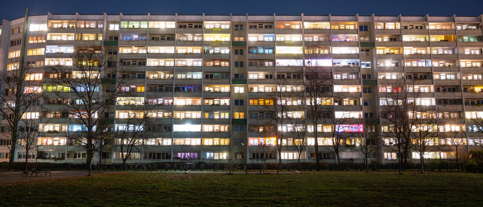 Ein mehrstöckiges Wohngebäude bei Nacht, dessen Fenster in verschiedenen Farben beleuchtet sind. Der Himmel ist klar und dunkel, und im Vordergrund ist eine Rasenfläche zu sehen. Einige Bäume stehen vor dem Gebäude. Die Szene vermittelt eine ruhige, nächtliche Atmosphäre.