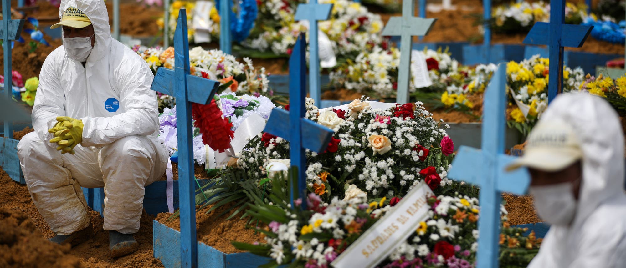 Friedhofsmitarbeiter in Schutzanzügen sitzen auf mit Blumen geschmückten Gräbern während einer Beerdigung am Friedhof Nossa Senhora Aparecida in Manaus.