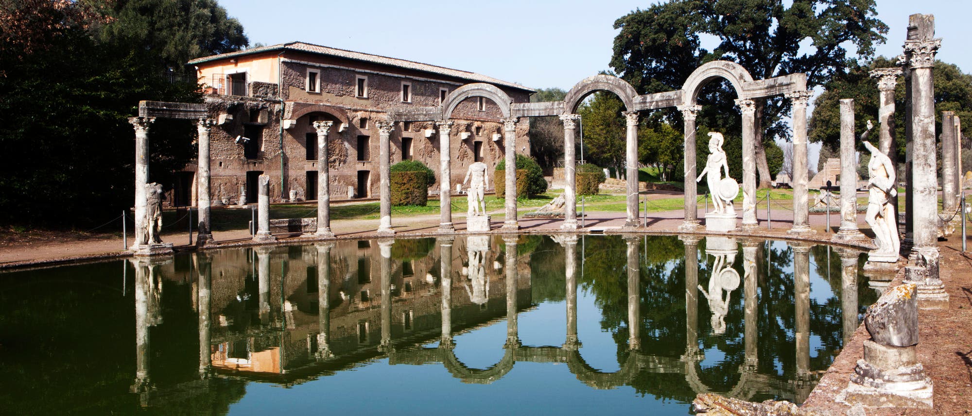 Eine antike römische Villa mit einem rechteckigen Wasserbecken im Vordergrund, umgeben von einer Kolonnade aus Säulen und Statuen. Die Reflexionen der Architektur und Skulpturen sind im ruhigen Wasser sichtbar. Im Hintergrund steht ein historisches Gebäude aus Stein, umgeben von Bäumen. Die Szene vermittelt eine ruhige und historische Atmosphäre. Eine antike römische Villa mit einem rechteckigen Wasserbecken im Vordergrund, umgeben von einer Kolonnade aus Säulen und Statuen. Die Reflexionen der Architektur und Skulpturen sind im ruhigen Wasser sichtbar. Im Hintergrund steht ein historisches Gebäude aus Stein, umgeben von Bäumen. Die Szene vermittelt eine ruhige und historische Atmosphäre.