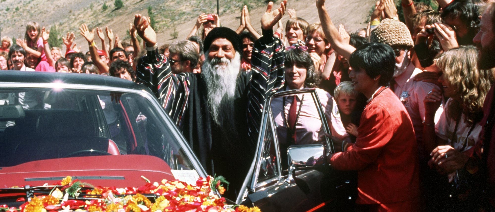 Eine Gruppe von Menschen steht um ein Auto, das mit Blumen bedeckt ist. Eine Person in der Mitte hebt die Arme, während die umstehenden Menschen klatschen und jubeln. Die Szene spielt sich in einer bergigen Landschaft ab. Die Stimmung wirkt feierlich und freudig.