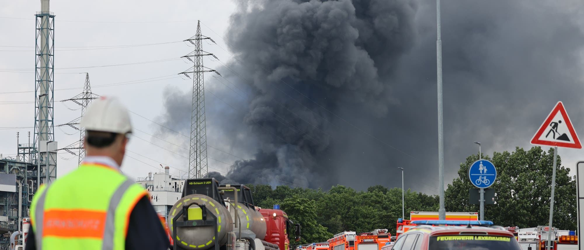 Einsatzfahrzeuge der Feuerwehr stehen am 27. Juli 2021 unweit einer Zufahrt zum Chempark, über dem eine dunkle Rauchwolke aufsteigt. 