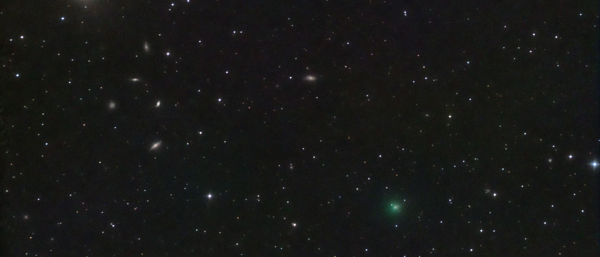 Ein astronomisches Bild zeigt einen sternen&uuml;bers&auml;ten Nachthimmel mit mehreren Galaxien. Links oben ist eine helle, elliptische Galaxie zu sehen, umgeben von zahlreichen kleineren, unscharfen Galaxien. &Uuml;ber das Bild verteilt sind viele leuchtende Sterne unterschiedlicher Helligkeit. In der unteren rechten Bildh&auml;lfte ist ein schwach gr&uuml;nlich leuchtendes Objekt zu erkennen, m&ouml;glicherweise ein Komet. Der Hintergrund ist tiefschwarz, was die Helligkeit der Himmelsk&ouml;rper hervorhebt.