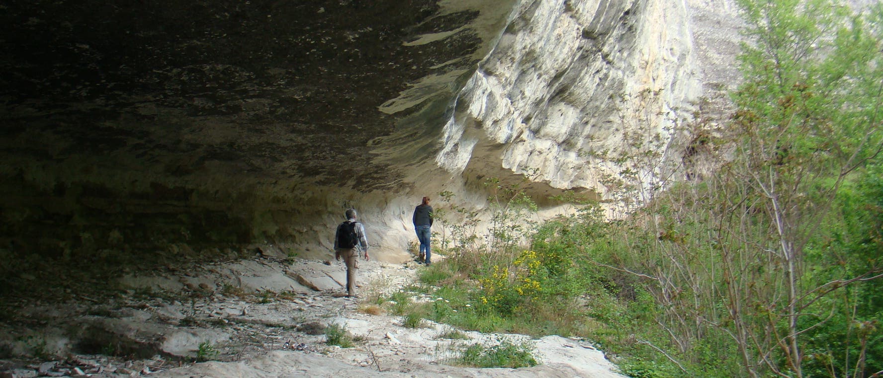 Zwei Personen wandern unter einem großen Felsüberhang in einer natürlichen Umgebung. Der Fels ist hell und von Vegetation umgeben. Die Szene vermittelt ein Gefühl von Abenteuer und Erkundung in der Natur.