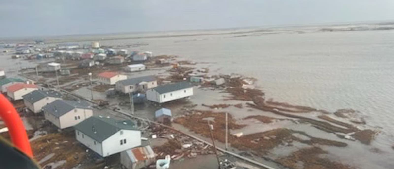 Luftaufnahme einer Küstenstadt, die von Überschwemmungen betroffen ist. Mehrere Häuser stehen in flachem Wasser, und die Straßen sind teilweise überflutet. Der Himmel ist bewölkt, was auf schlechtes Wetter hindeutet. Die Szene zeigt die Auswirkungen von Hochwasser auf die Infrastruktur der Stadt.