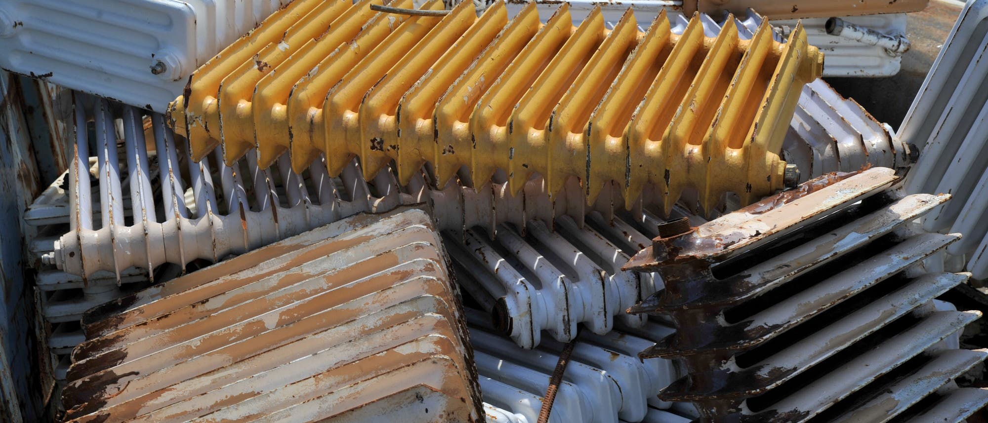 Ein Stapel alter, rostiger Heizk&ouml;rper in verschiedenen Farben, darunter Wei&szlig;, Gelb und Braun. Die Heizk&ouml;rper sind ungleichm&auml;&szlig;ig gestapelt und zeigen deutliche Gebrauchsspuren und Roststellen. Die Szene vermittelt den Eindruck eines Schrottplatzes oder einer Sammlung von Altmetall.