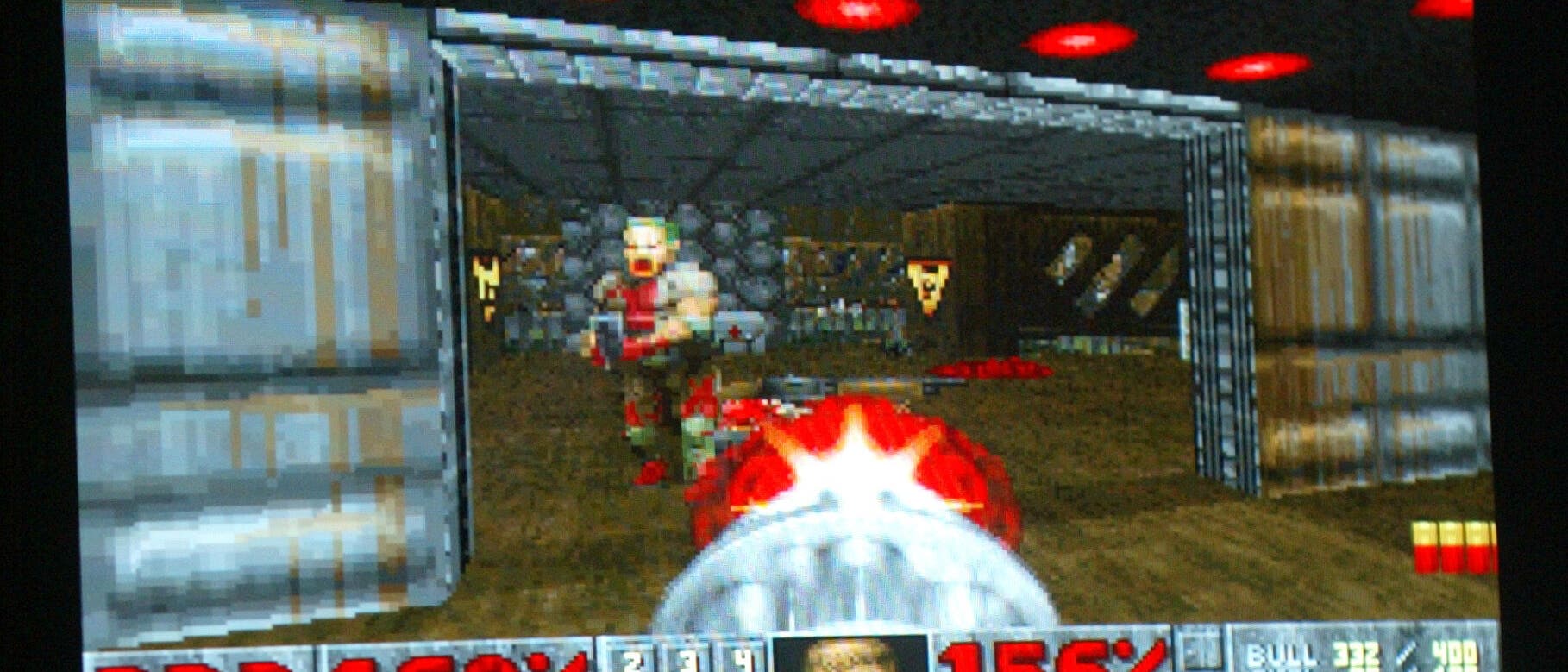 Ein Bildschirmfoto eines klassischen Egoshooter-Videospiels, das auf einem Monitor angezeigt wird. Im Vordergrund ist eine Hand auf einer Tastatur zu sehen, die das Spiel steuert. Auf dem Bildschirm ist ein Spielcharakter in einer futuristischen Umgebung zu erkennen, der eine Waffe abfeuert. Unten auf dem Bildschirm sind Statusanzeigen für Munition, Gesundheit und Rüstung zu sehen. Das Bild vermittelt eine aktive Spielszene.