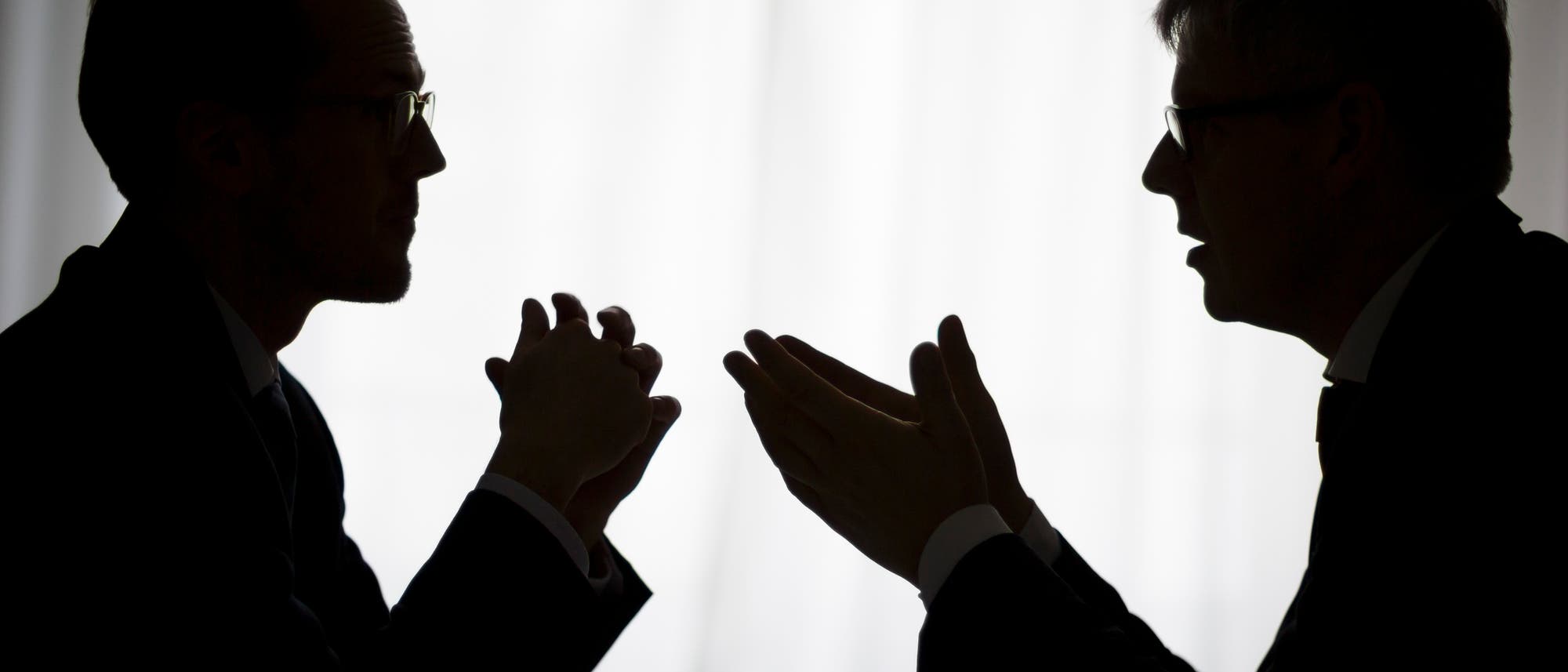 Silhouette zweier gestikulierender Männer im Gespräch vor hellem Hintergrund