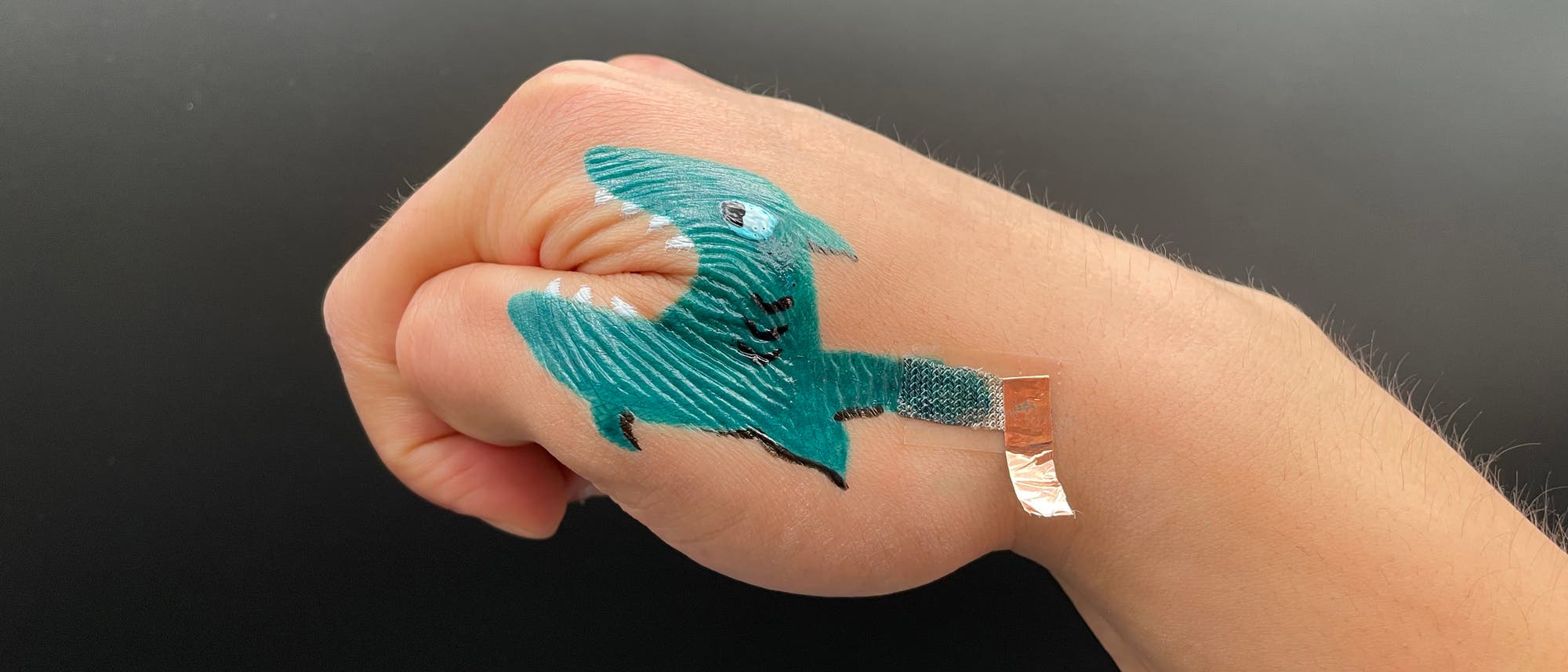 Eine Hand mit einer künstlerischen Zeichnung eines Haifischs, die über die Finger und den Handrücken verläuft. Der Mund des Haifischs ist so positioniert, dass er sich öffnet, wenn die Hand geöffnet wird. Ein kleines Stück Metallfolie ist an der Zeichnung befestigt. Eine Hand mit einer künstlerischen Zeichnung eines Haifischs, die über die Finger und den Handrücken verläuft. Der Mund des Haifischs ist so positioniert, dass er sich öffnet, wenn die Hand geöffnet wird. Ein kleines Stück Metallfolie ist an der Zeichnung befestigt.