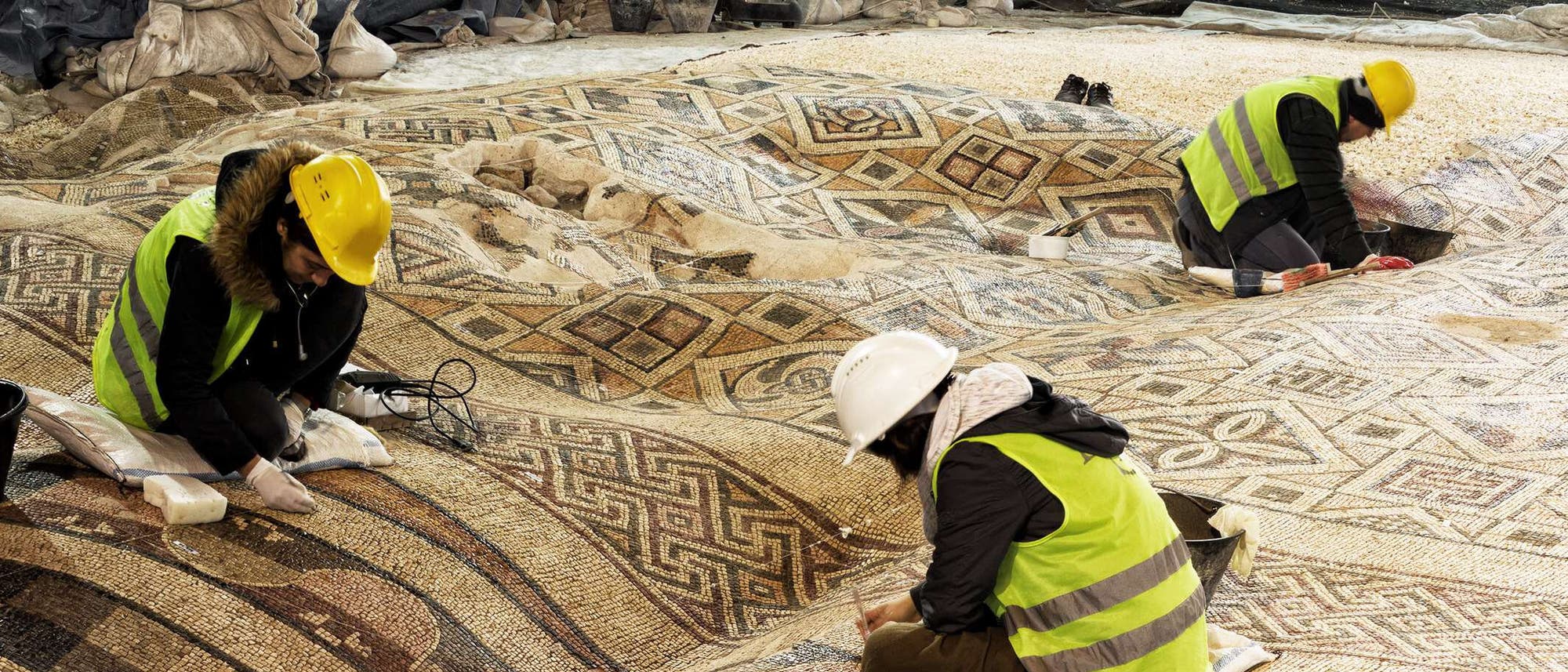 Drei Personen in Schutzkleidung und Helmen arbeiten an der Restaurierung eines gro&szlig;en, antiken Mosaiks mit geometrischen Mustern. Sie sind auf einem arch&auml;ologischen Ausgrabungsort besch&auml;ftigt und verwenden Werkzeuge zur Reinigung und Konservierung der Mosaiksteine. Die Szene zeigt die Detailarbeit, die in die Erhaltung historischer Kunstwerke investiert wird.