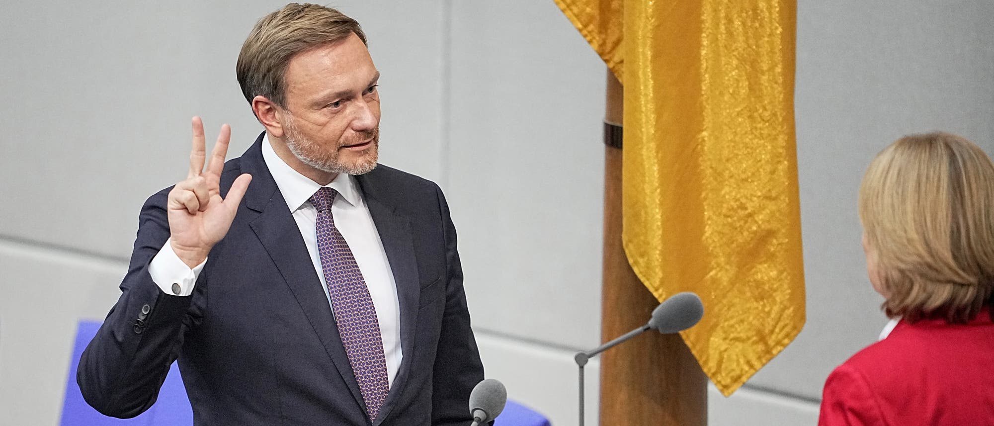 FDP-Politiker Christian Lindner steht im Plenarsaal des Deutschen Bundestags und hebt die rechte Hand, an der Daumen, Zeige- und Mittelfinger weggespreizt sind. Er trägt Anzug und Krawatte. 