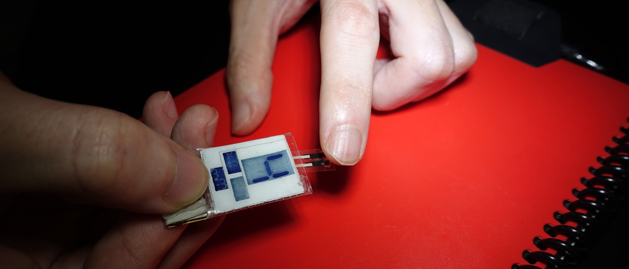 Eine Hand hält einen improvisiert anmutenden Biosensor mit LCD-Nummernanzeige gegen eine Fingerspitze.