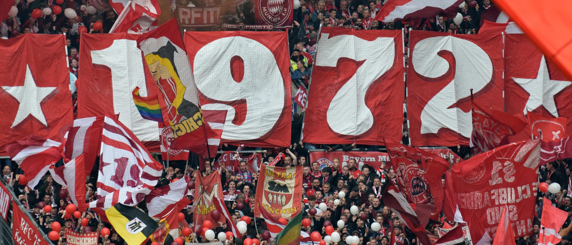Eine große Menschenmenge in einem Fußballstadion hält rote Banner mit der weißen Aufschrift »1972« hoch. Um die Zahlen herum sind rote und weiße Fahnen und Schals zu sehen. Die Fans jubeln und schwenken rote und weiße Luftballons. Im Hintergrund sind weitere Fans und Fahnen zu erkennen. Eine große Menschenmenge in einem Fußballstadion hält rote Banner mit der weißen Aufschrift »1972« hoch. Um die Zahlen herum sind rote und weiße Fahnen und Schals zu sehen. Die Fans jubeln und schwenken rote und weiße Luftballons. Im Hintergrund sind weitere Fans und Fahnen zu erkennen.