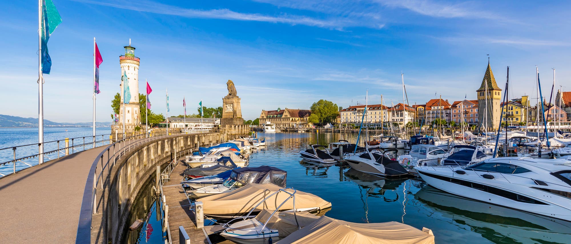 Hafen von Lindau am Bodensee bei sonnigem Wetter. Links ist die Hafenmole mit dem markanten Leuchtturm und der Löwenstatue zu sehen. Im Vordergrund liegen Boote am Steg, einige mit Abdeckungen. Rechts erstreckt sich die Altstadt mit historischen Gebäuden und einem Kirchturm. Der Himmel ist klar und blau.