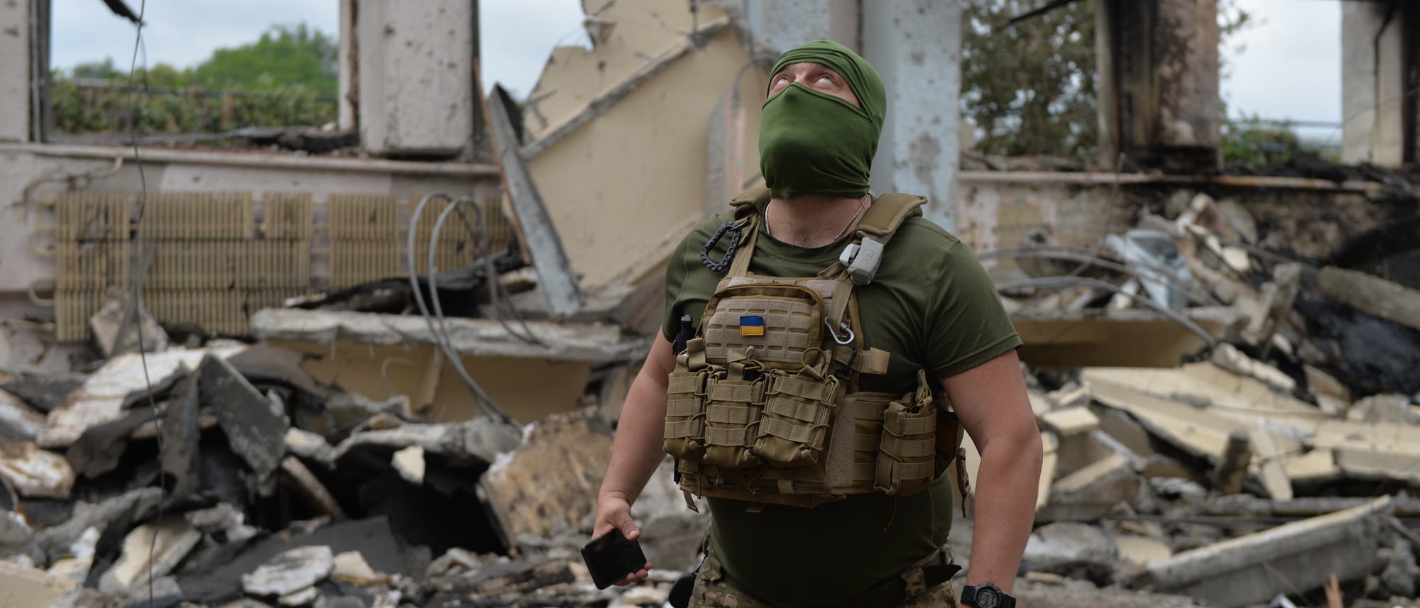 Ein mit grüner Maske vermummter Soldat schaut zur Decke. Er steht inmitten der Trümmer eines Gebäudes.