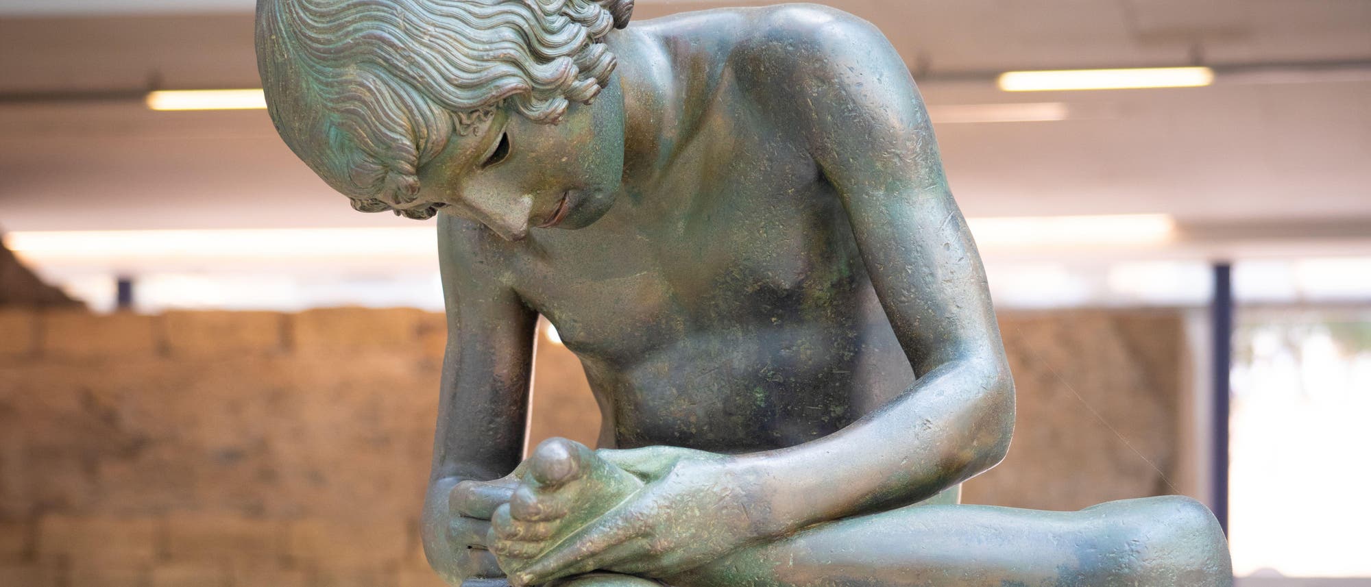 Eine Bronzestatue eines jungen Mannes, der auf einem Felsen sitzt und einen Dorn aus seinem Fu&szlig; zieht. Die Skulptur befindet sich in einem Museum mit Steinw&auml;nden und einer gro&szlig;en Fensterfront im Hintergrund. Der Raum ist gut beleuchtet und die Statue steht auf einem runden Sockel.