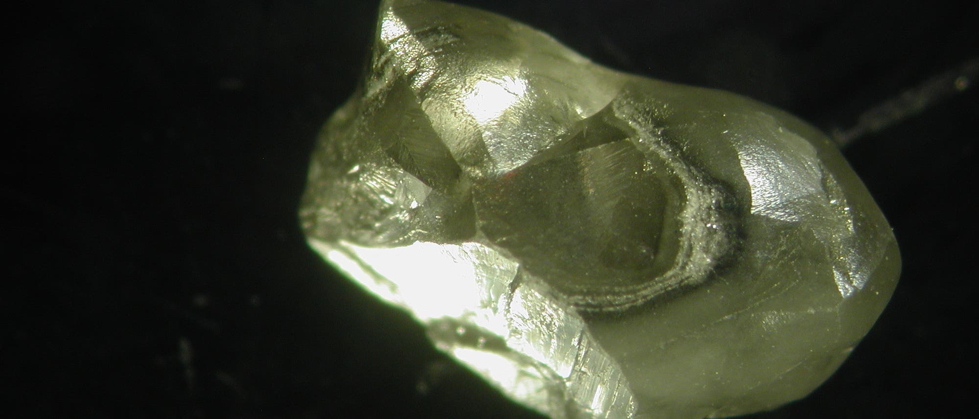 Nahaufnahme eines rohen, ungeschliffenen Diamanten auf schwarzem Hintergrund. Der Diamant zeigt eine unregelmäßige, kristalline Struktur mit glänzenden, reflektierenden Flächen. Die Lichtreflexionen betonen die Transparenz und die natürliche Form des Steins.