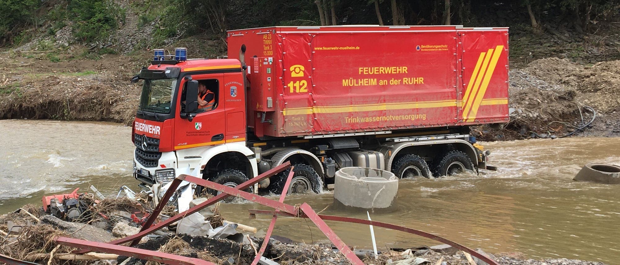 Ein Feuerwehrfahrzeug der Feuerwehr Mülheim an der Ruhr fährt durch ein überflutetes Gebiet. Auf dem roten Fahrzeug sind die Aufschrift "Trinkwasserversorgung" und die Notrufnummer 112 zu sehen. Im Vordergrund liegen Trümmerteile im Wasser. Im Hintergrund sind Bäume und ein schlammiger Hang zu erkennen.