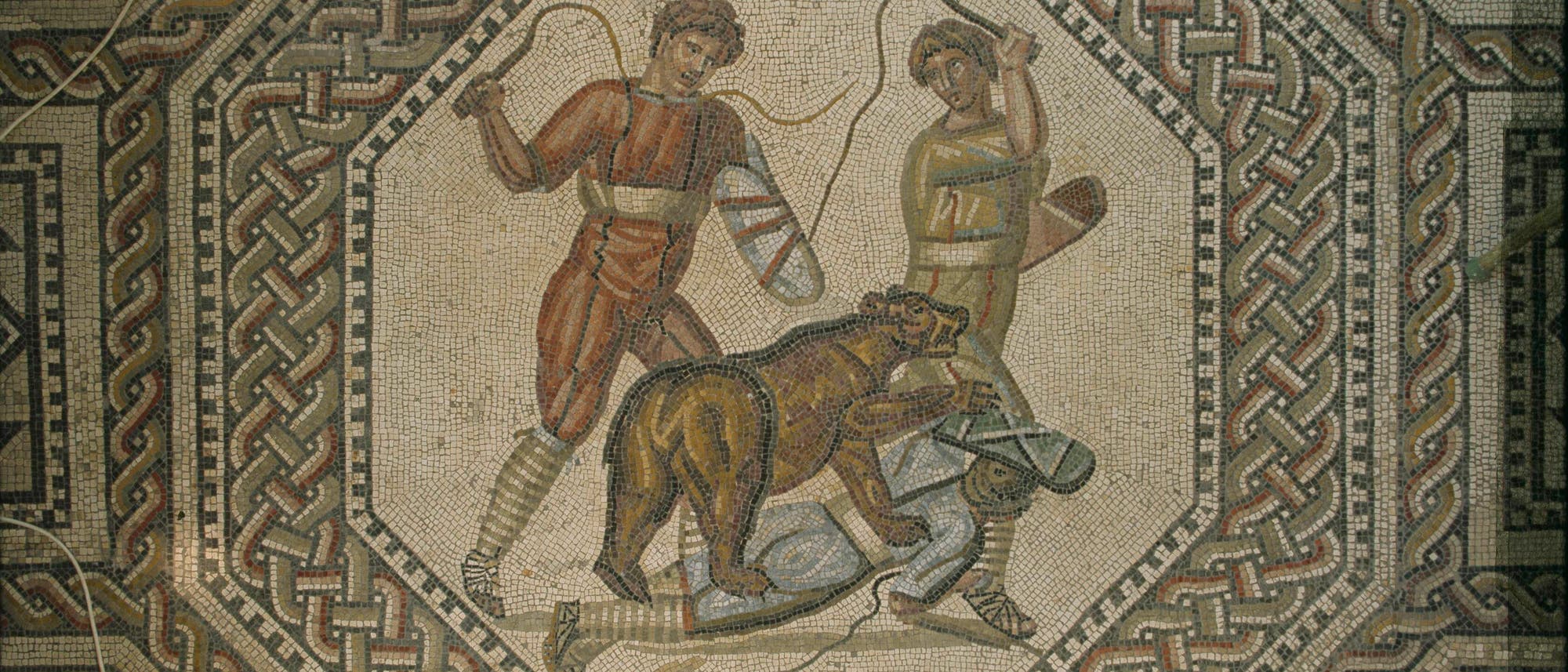 Ein antikes Mosaik zeigt zwei Personen in römischer Kleidung, die mit Peitschen gegen einen Bären vorgehen. Der Bär steht über einer dritten Person, die am Boden liegt. Das Mosaik ist von einem komplexen geometrischen Muster umrahmt, das aus ineinander verschlungenen Bändern besteht. Die Szene vermittelt eine dynamische Darstellung von Bewegung und Spannung.