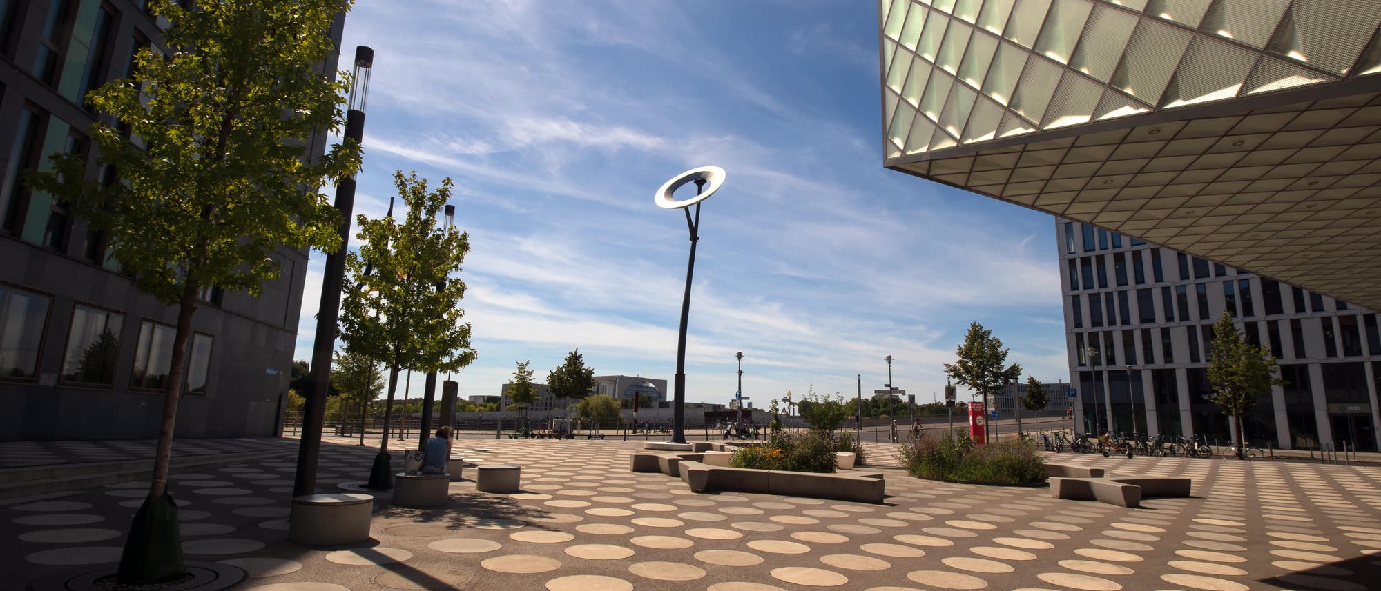 Vor einem fast klaren blauen Himmel auf einem öffentlichen Platz steht ein Kunstwerk, das aus einem 15 Meter hohen Metallpfahl besteht und einer an dessen Spitze aufgehängten, in der Sonne glänzenden Metallscheibe, deren Durchmesser etwa vier Meter beträgt.