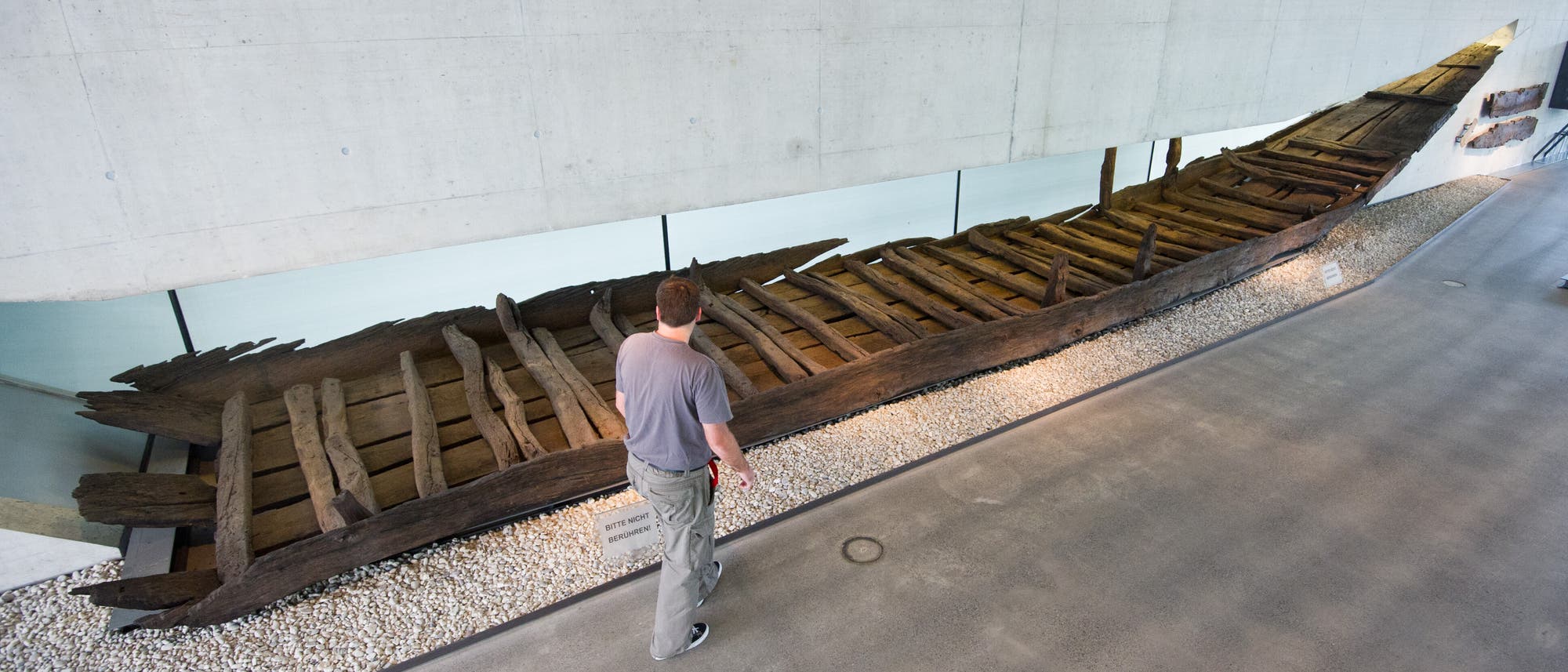 Eine Person betrachtet ein antikes Holzboot, das in einem modernen Museum ausgestellt ist. Das Boot ist aus dunklem Holz gefertigt und zeigt deutliche Spuren von Alterung. Es liegt auf einem Bett aus kleinen weißen Steinen. Die Umgebung ist minimalistisch gestaltet, mit Betonwänden und einer Glasbarriere, die das Exponat schützt. Ein Schild mit Informationen ist neben dem Boot platziert. Eine Person betrachtet ein antikes Holzboot, das in einem modernen Museum ausgestellt ist. Das Boot ist aus dunklem Holz gefertigt und zeigt deutliche Spuren von Alterung. Es liegt auf einem Bett aus kleinen weißen Steinen. Die Umgebung ist minimalistisch gestaltet, mit Betonwänden und einer Glasbarriere, die das Exponat schützt. Ein Schild mit Informationen ist neben dem Boot platziert.