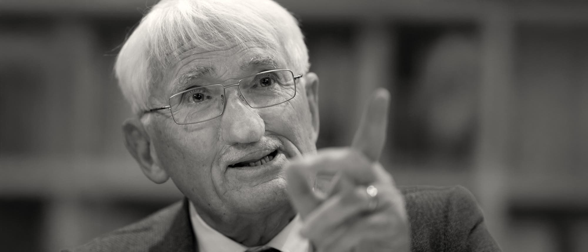 Ein &auml;lterer Mann mit wei&szlig;en Haaren und Brille schaut nach oben leicht an der Kamera vorbei. Er hebt den Zeigefinger seiner linken Hand und scheint zu mahnen oder zu diskutieren.