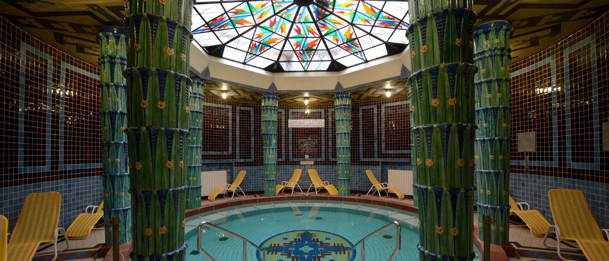  Ein kunstvoll gestalteter Innenraum eines Badehauses mit einem runden Pool in der Mitte, umgeben von gr&uuml;nen, verzierten S&auml;ulen. Der Boden des Pools zeigt ein geometrisches Mosaikmuster. &Uuml;ber dem Pool befindet sich eine bunte, glasierte Kuppel, die das Licht in verschiedenen Farben bricht. Um den Pool herum stehen gelbe Liegest&uuml;hle. Die W&auml;nde sind mit blauen Fliesen verkleidet, die ein harmonisches Gesamtbild schaffen. 