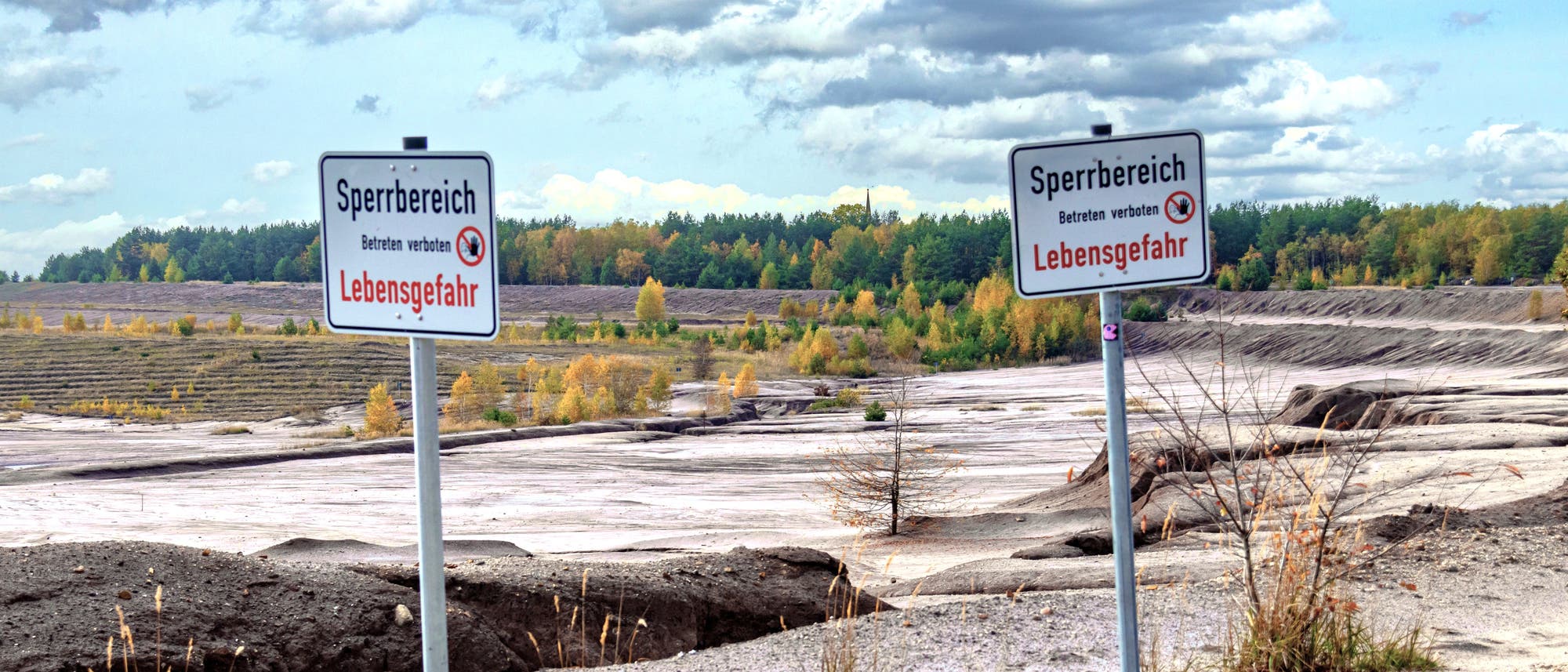 Warnschilder im Vordergrund, dahinter karge Sandlandschaft mit Erosionsrinnen, im Hintergrund ein Wald